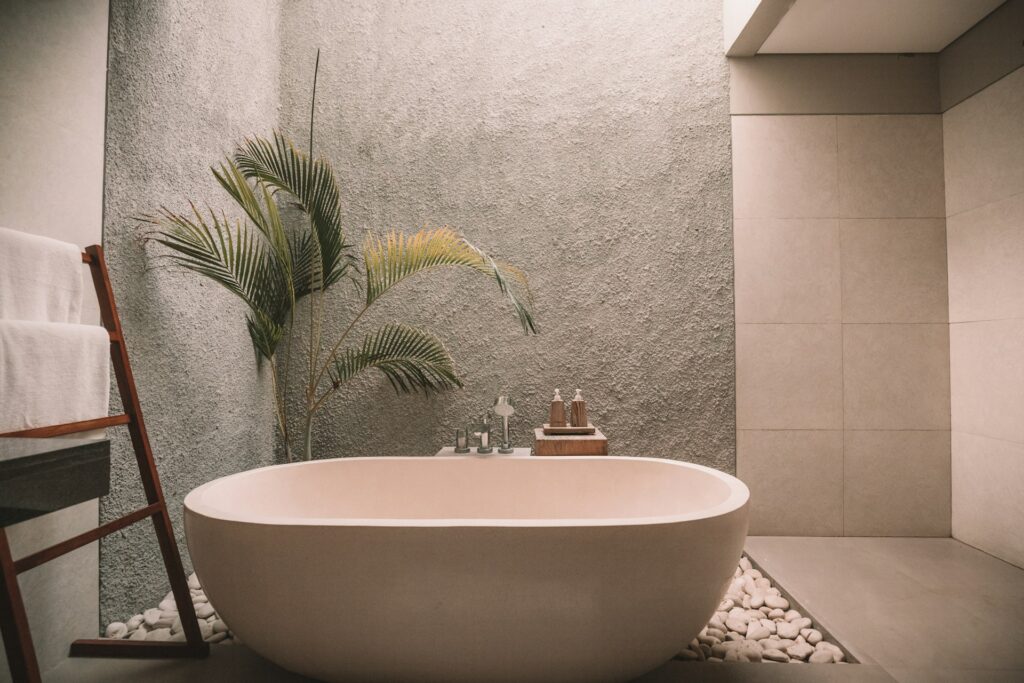 A white stand-alone tub sits in a bathroom made almost entirely of white tiles. Behind the tub is a floor of white rocks. The corner is made of exposed concrete walls. A large plant sits behind the tub underneath bright sunlight, likely from a skylight.