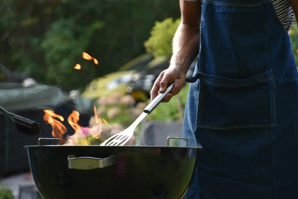 A person wearing a jean apron stands over a small grill with a stainless steel spatula. Flames leap from the grill. Trees are in the background.