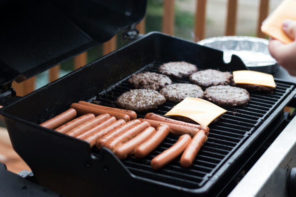 Burgers and hot dogs sit on a grill with a low flame. Two burgers also have two pieces of cheese. A person stands by the grill with more cheese for the remaining burgers.