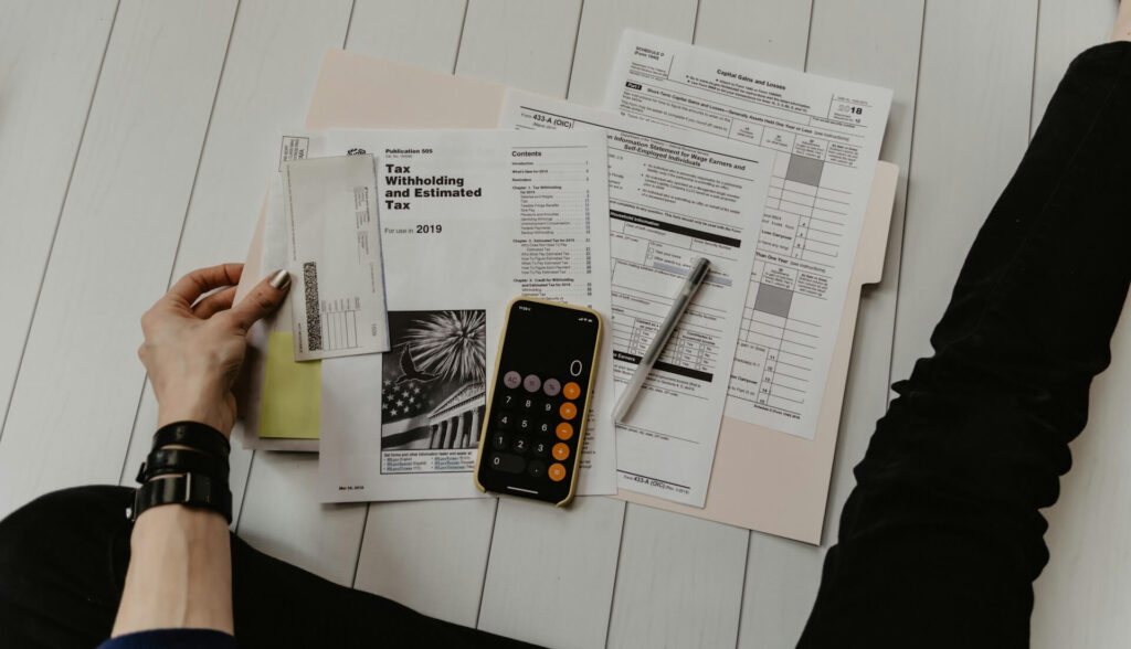 A person sitting on a white floor made with thin wooden boards spreads their estimated tax papers out on top of a light pink folder. An iPhone in a yellow case displaying the calculator app and a black ink pen sit on top of the papers. The person wears black jeans and black bracelets.