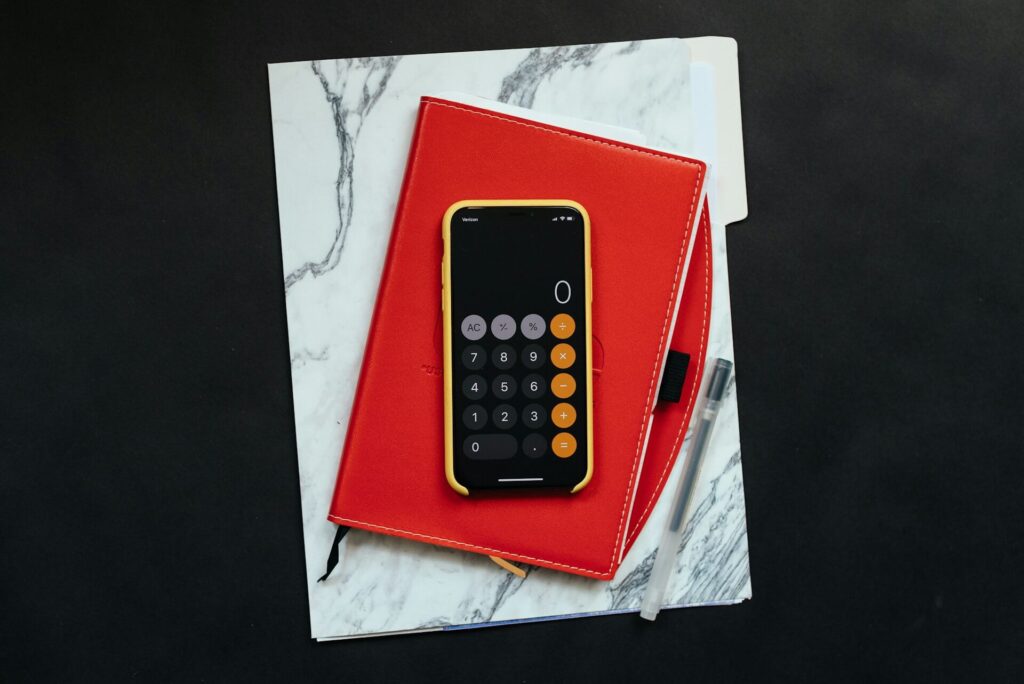 A marble folder, red planner and iPhone in a yellow case displaying the calculator app sit on top of a black surface. A black ink pen sits on top of the marble folder.