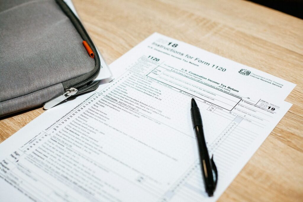 Two pages for an IRS Income Tax 1120 Form sit on a light wooden desk next to a zippered gray bag containing other papers. A black pen sits on top of the 1120 forms.