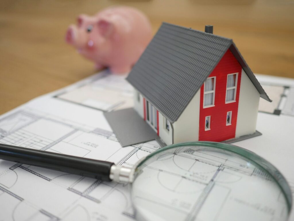 A miniature red and white house sits on a printed page of floor plans. A magnifier glass and a pink piggy bank sit next to it.