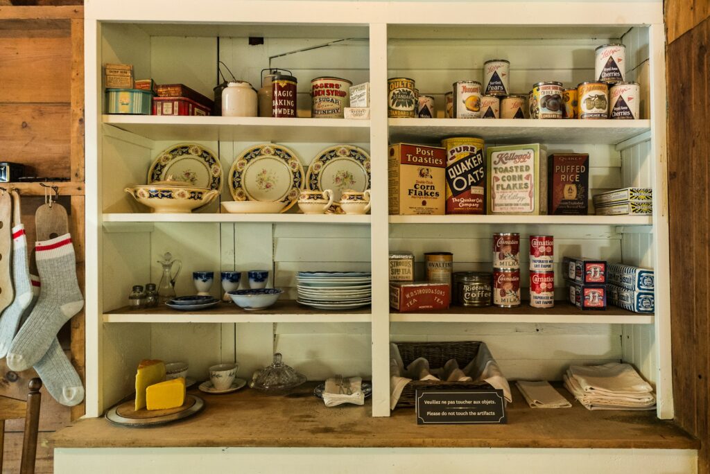 A white freestanding kitchen pantry stands against a wooden wall. It has dishes and groceries, plus a decorative sign on the wooden shelf in the middle.