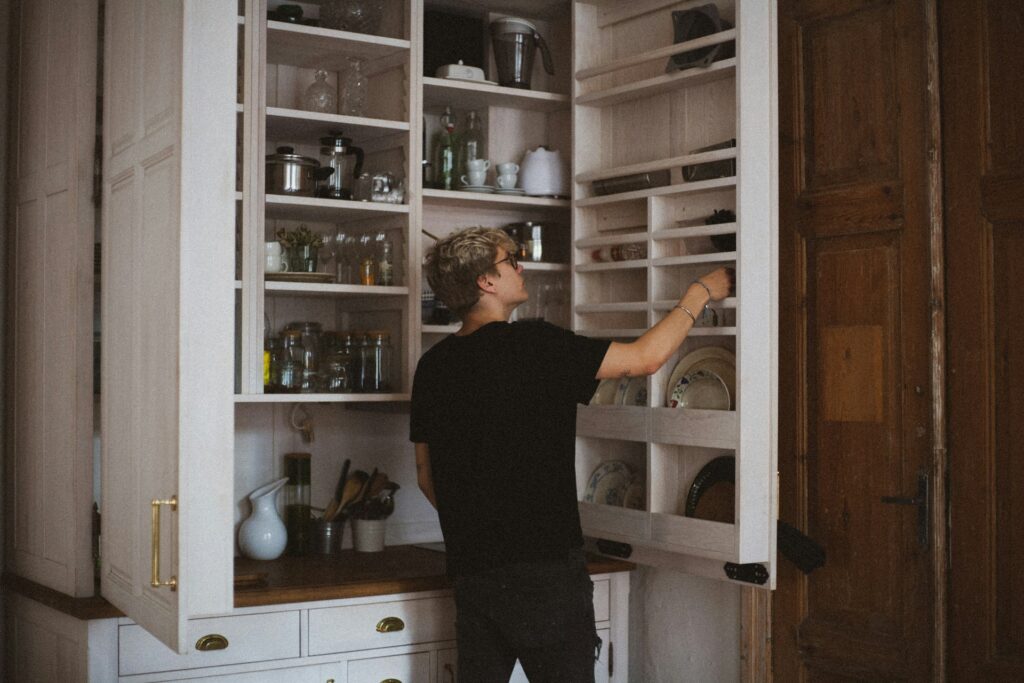 A person with short blond hair and an undercut opens the shelving of a pantry. It's white and has a brown counter underneath. Beneath the counter are three drawers and more cabinets.