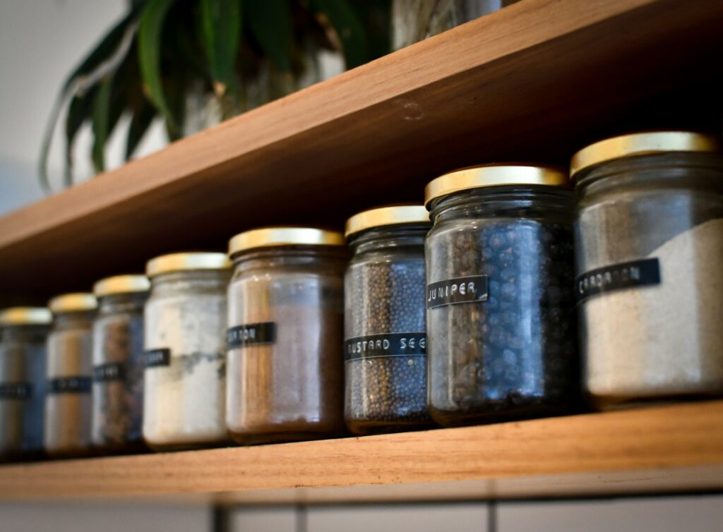 Two light wooden shelves sit on a white wall. They contain mason jars with golden lids and typewriter letter labels for the spices within each one. A green plant sits on the top shelf.