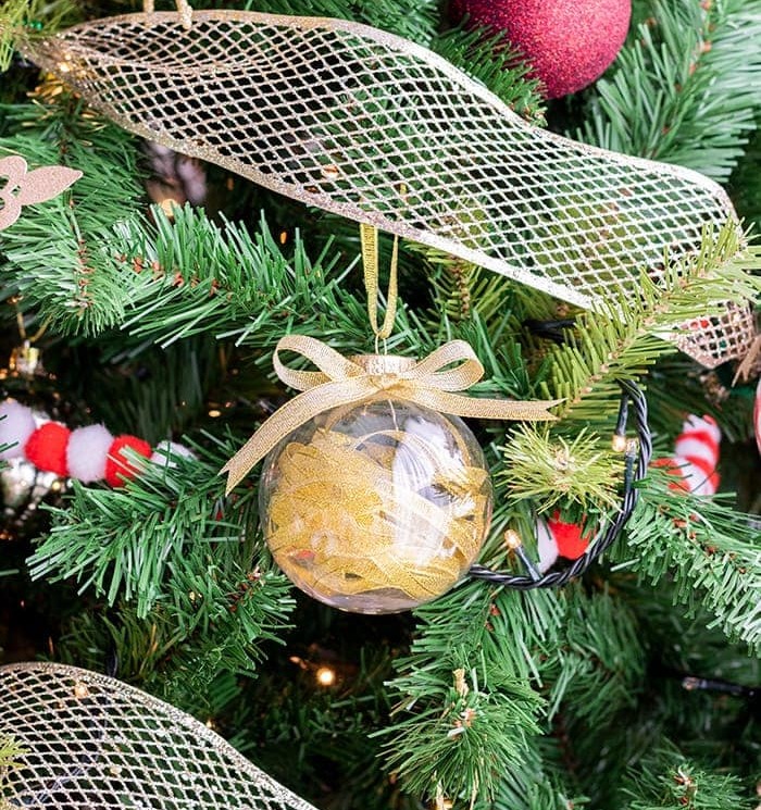 A glass ornament filled with yellow ribbons hangs from a gold ribbon on a fake fir Christmas tree among red ornaments, mesh ribbons and yellow-white string lights.