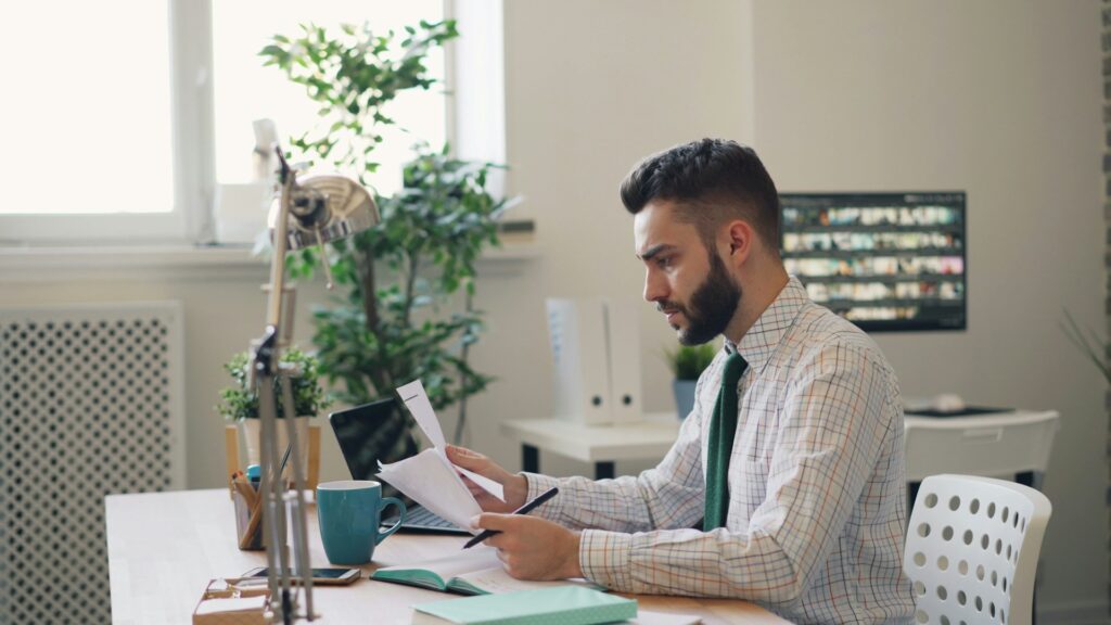 A guy in a thin plaid shirt and green tie reviews paperwork at his professional desk. His desk is covered in notebooks, plants, a laptop, a coffee mug and his phone. Another desk sits in the background, which makes this presumably a rental office where people would go to break their apartment lease.
