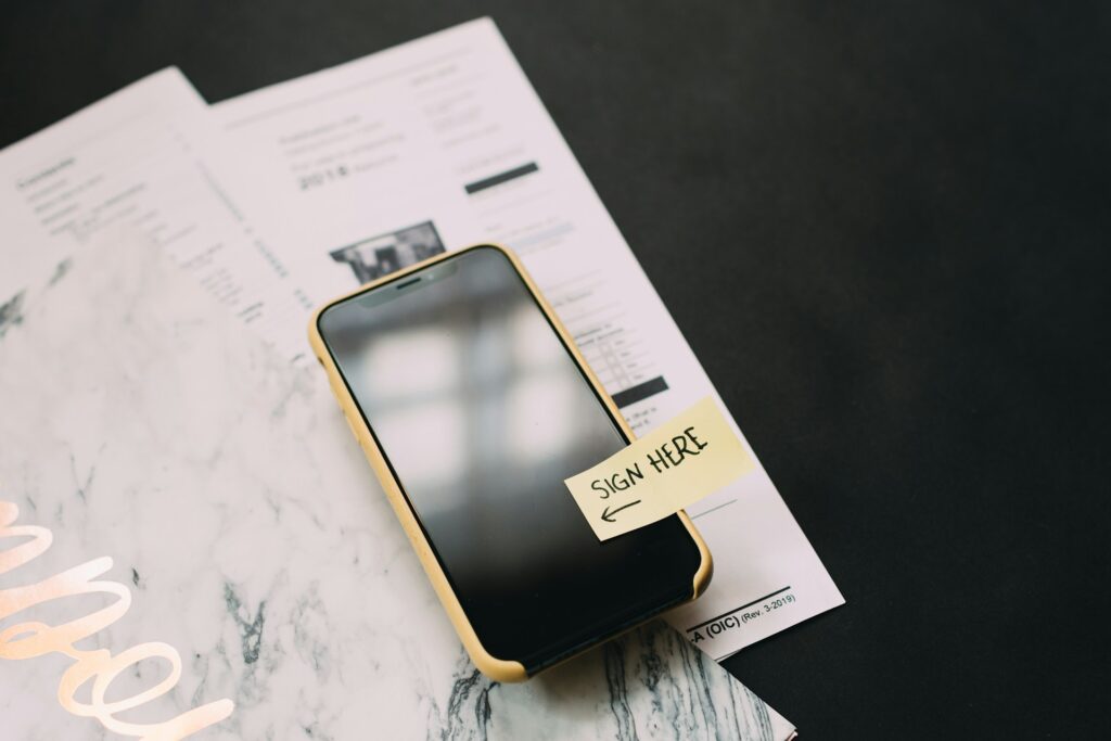 An iPhone in a yellow case has a yellow sticky tab on the screen. The tab says "Sign Here." It's on top of paperwork on a black surface.
