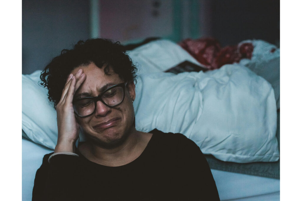 A woman grimacing and touching her forehead while sitting on the floor and leaning against the bed.