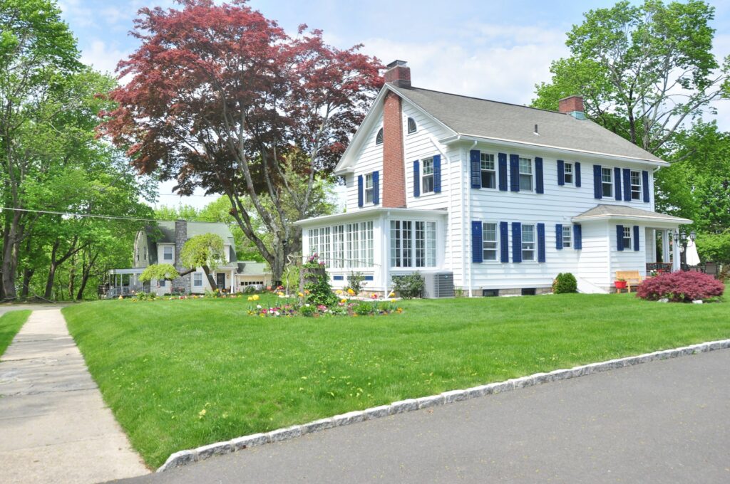 A two-story white house with navy shutters and a glass sun room sits among green and red trees. It has a lush green lawn and blooming flowerbeds. The sky is blue with some clouds.