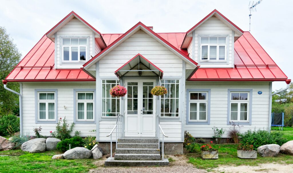 A small white wooden house with a red roof sits on an exposed stone foundation. The sky is cloudy above it and bright green grass with flowers grow around the house.