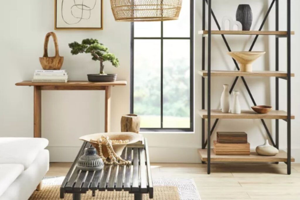 A modern living room with a white sofa, wooden tables with decorative objects, a bookshelf, and a large window letting in natural light.