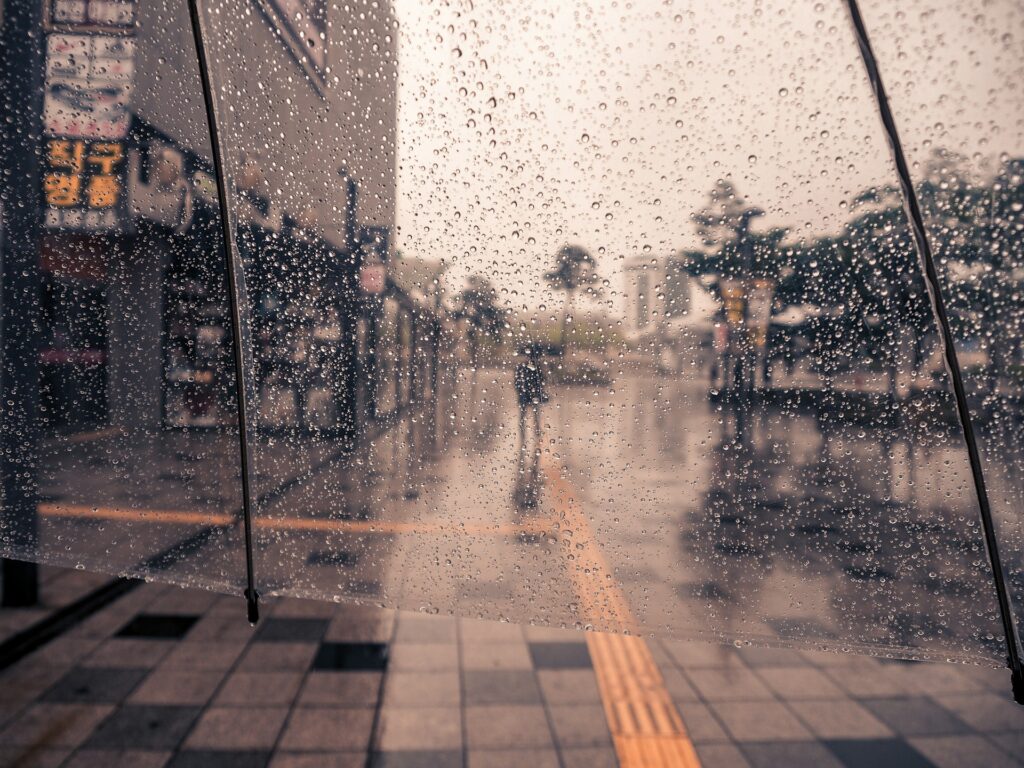 Raindrops sit on a clear umbrella under a gray sky. People walk on the wet city street beyond it.