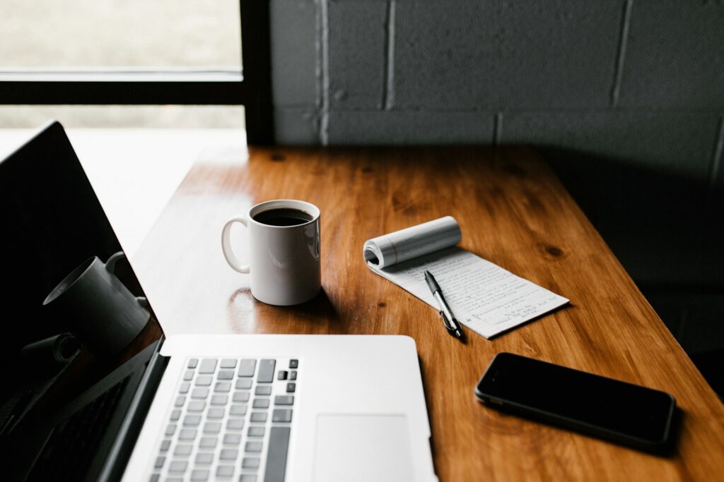 A laptop, phone, mug, pad of paper and pen on a wooden desk