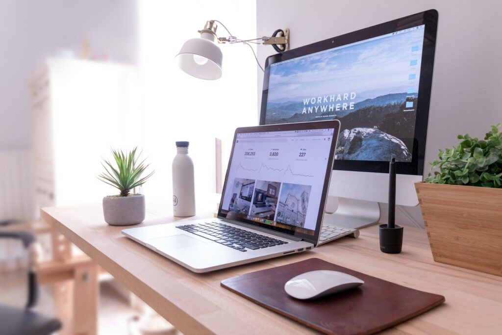 A work-from-home office with a laptop, monitor, mouse, water bottle and two plants on a wooden desk