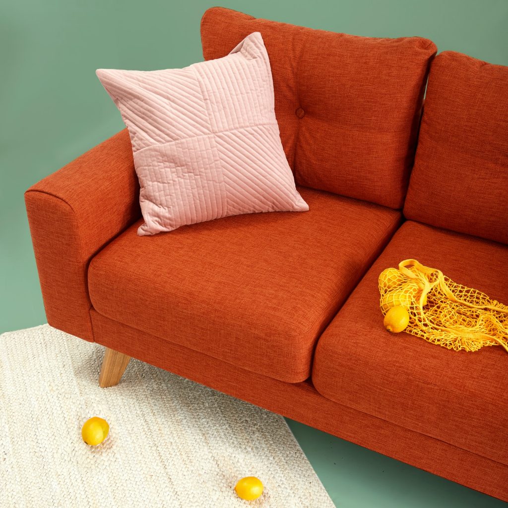 Orange sofa on a beige woven rug with a pastel pink pillow and a yellow bag of lemons on it