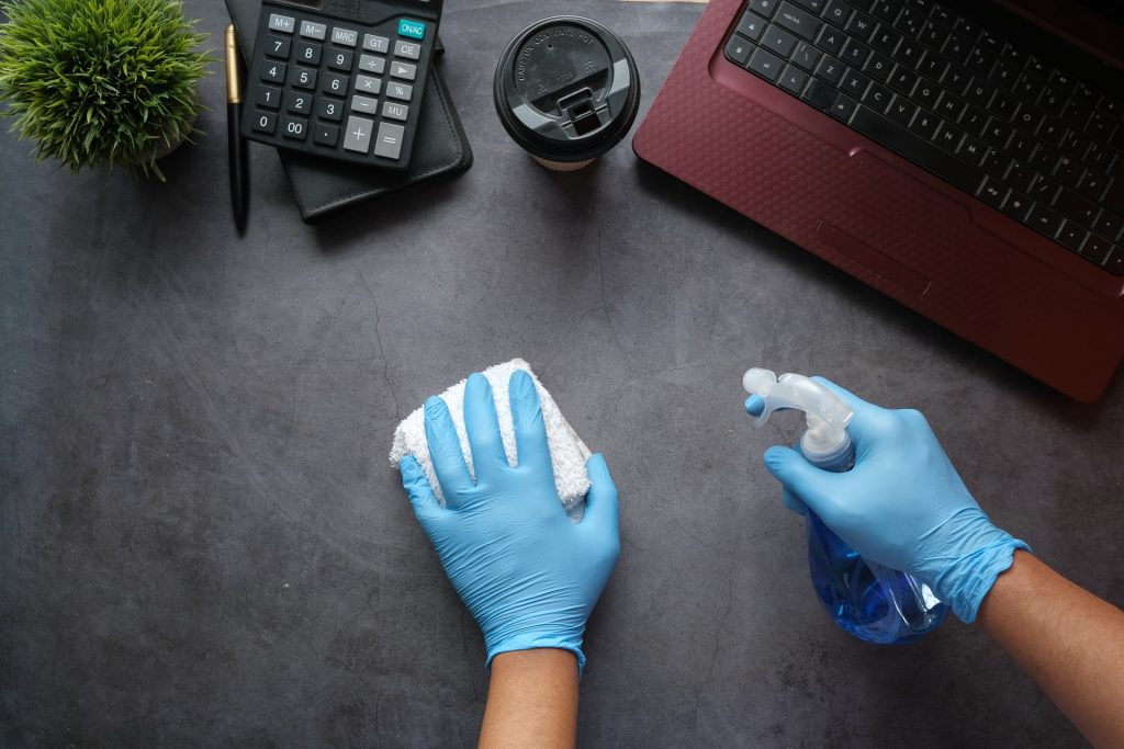 A person with blue latex gloves spraying and wiping a grey desk