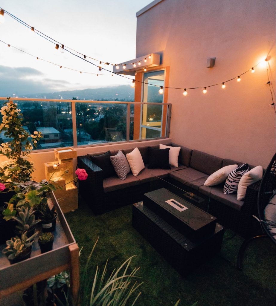 A small, turfed yard with string lights, plants, a firepit, and a couch