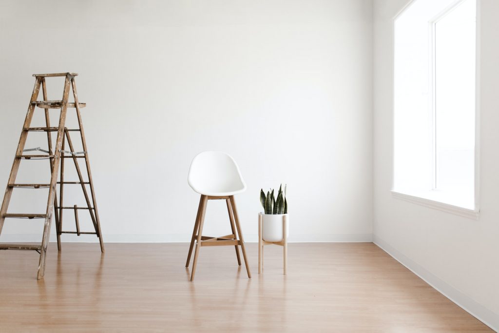 A wooden ladder in a bare white room witha white chair and a houseplant