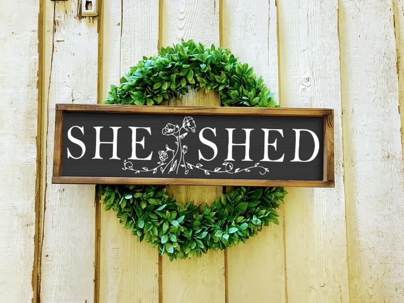 a sign with she shed written on it against a greenery wreath