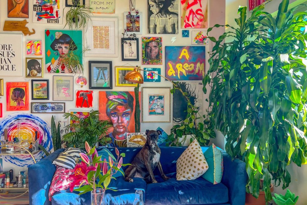 An apartment wall covered in tons of brightly colored artwork. A large green plant covers almost all of the window. There is a blue couch with a gray dog sitting on it and pink flowers in a vase.