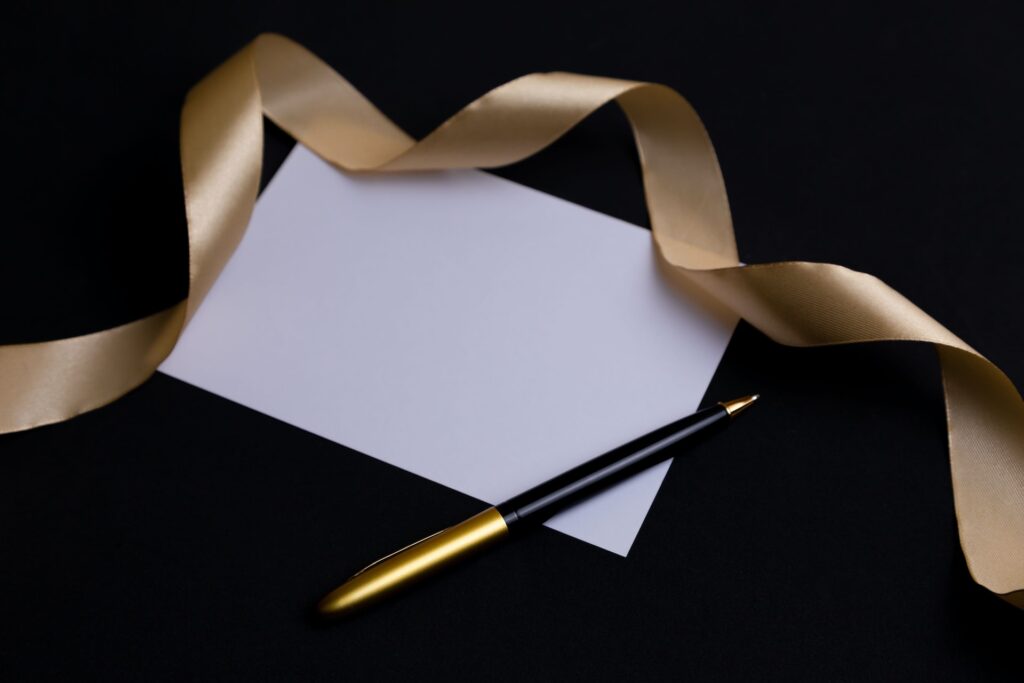 a blank white card against a black background with a gold ribbon and a black pen