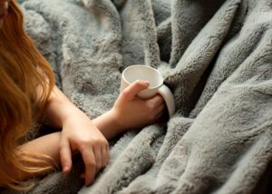 a woman with a steaming mug snuggled up with one of the best blankets for winter