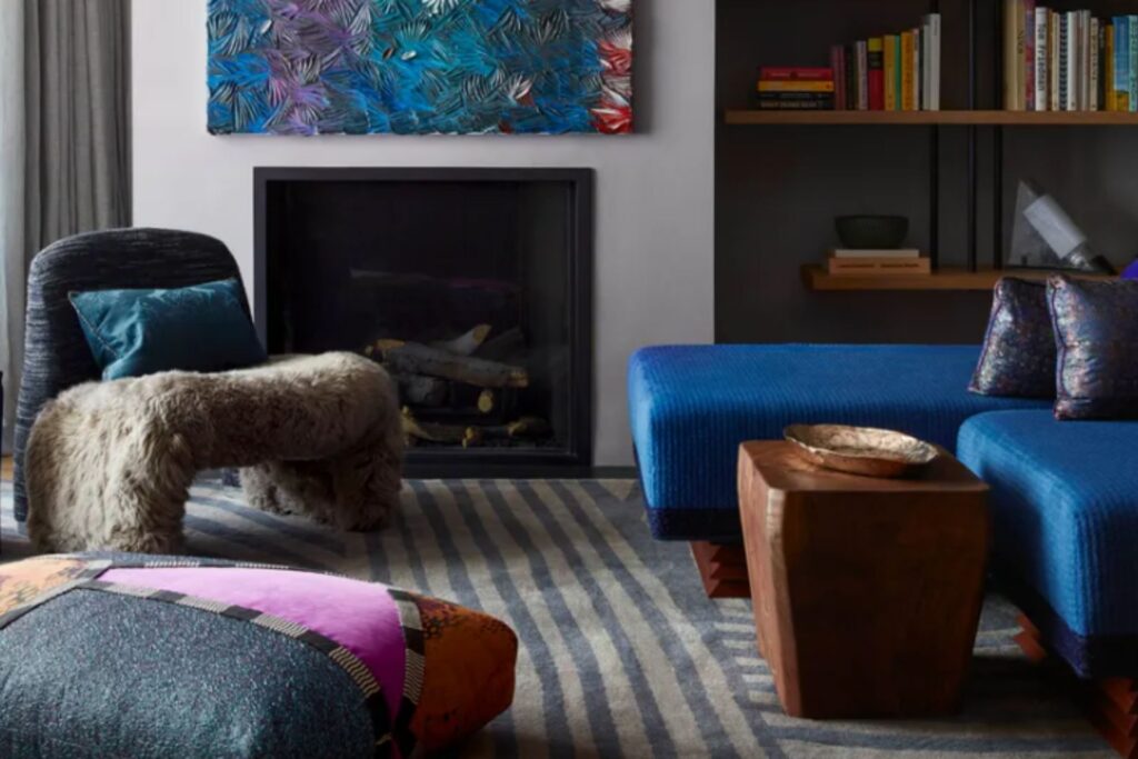 A cozy living room with a modern fireplace, a plush dark blue sofa with patterned pillows, a furry bench, a blue upholstered armchair with a dark teal cushion, and striped flooring.