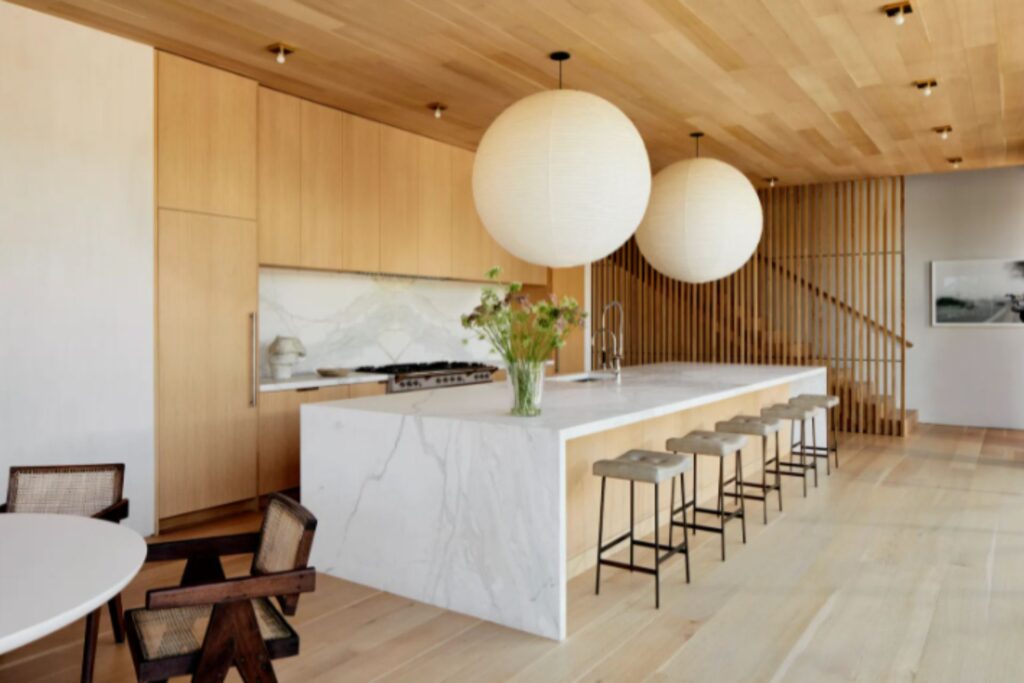A modern kitchen with light wood cabinets and ceiling, white marble island, wooden stools, and two large white pendant lights.