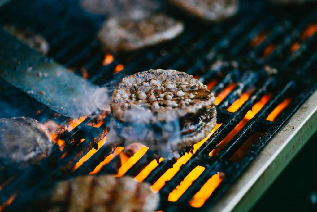 A person holds a stainless steel spatula near a burger that's on a grill. Other burgers sit around it. A fire goes beneath it. The grate is dirty, which is why it's important for the reader to learn how to clean gas grill grates.