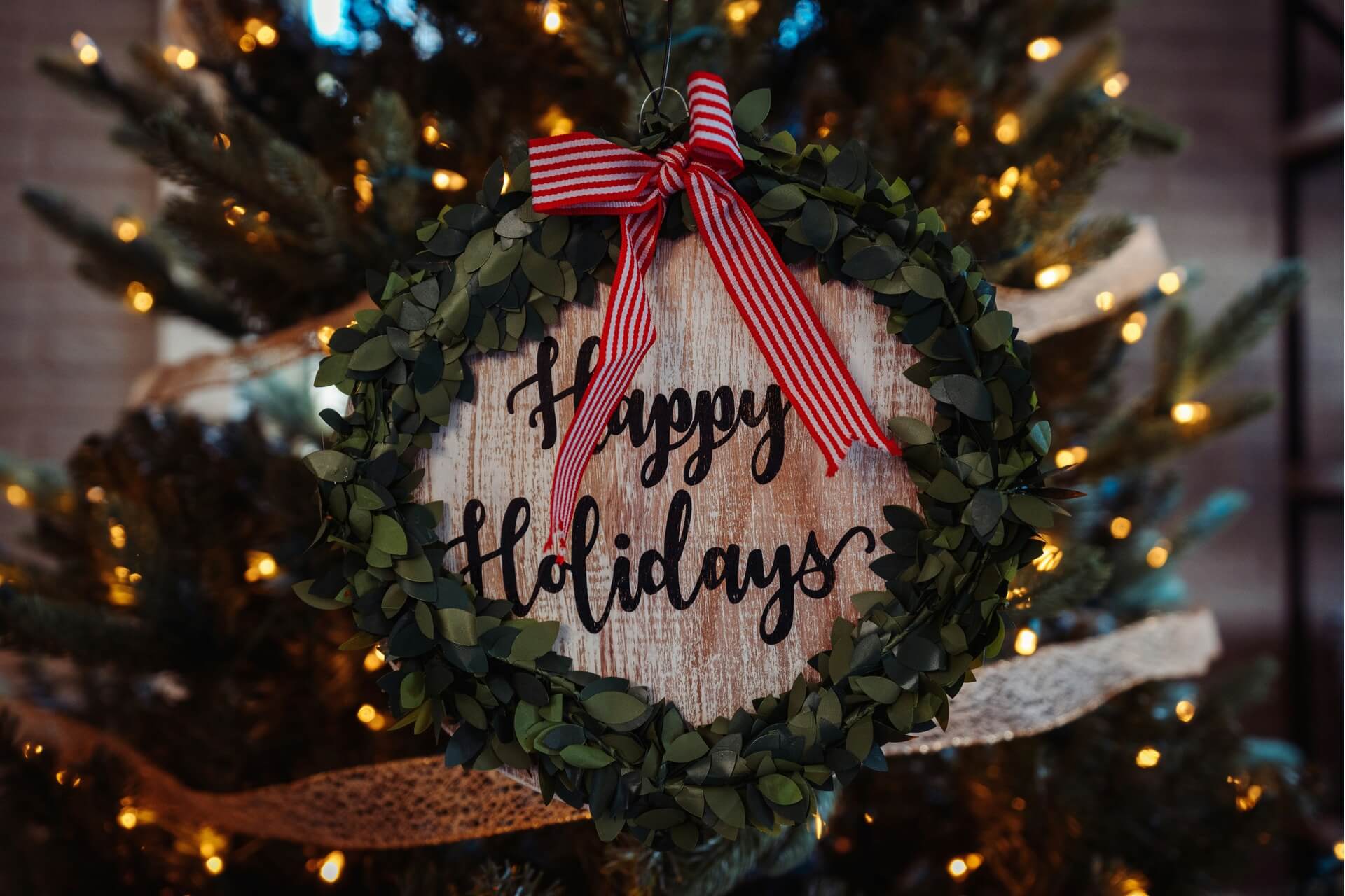 A Happy Holidays wooden board with a white and red ribbon and trimmed with leaves.