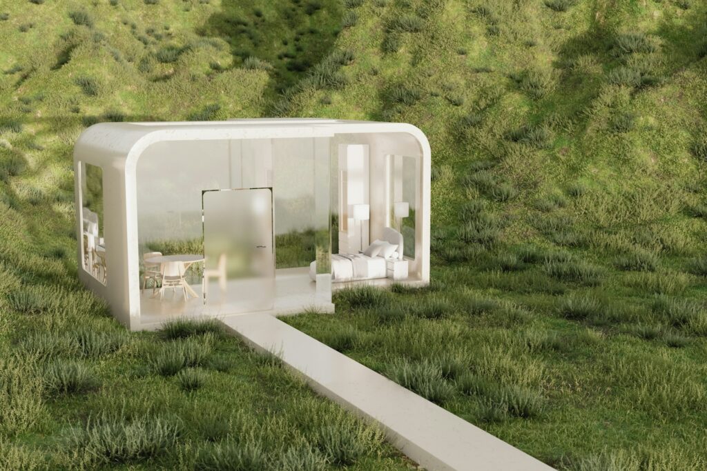 A tiny home with fogged windows sits on a grassy field at the base of a grassy hill. It has a white boarder and houses a kitchen and bedroom with minimal vertical storage.