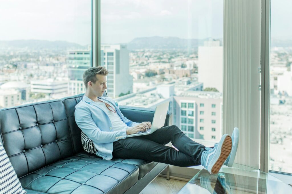 A man in a blue suit jacket, nice pants and light blue shoes sits on a leather couch. The walls behind him are floor-to-ceiling windows in a condo high above a city. He's typing on a Macbook, presumably looking at Amazon tiny houses.