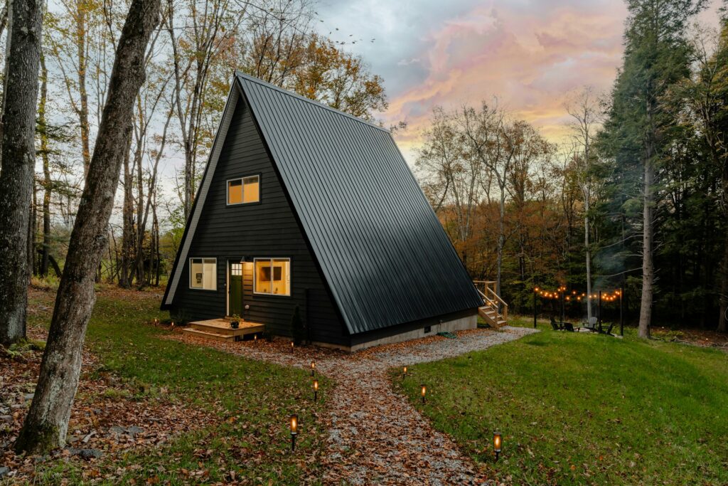 A tiny black A-frame home sits in a green yard amid trees on a mountainside. A campfire is going among some black chairs just off the back porch. String lights glow above the seating.