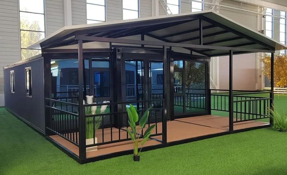 A black and metal luxury foldable Amazon tiny house sits on faux grass inside a warehouse. Potted plants are on and off the porch. The front wall has multiple doors and floor-to-ceiling windows.