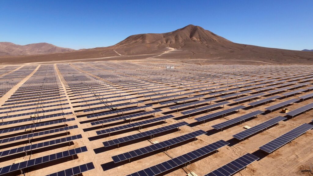 A field of solar panels sits in a sandy desert with rocky, plant-less hills in the background under a blue sky.