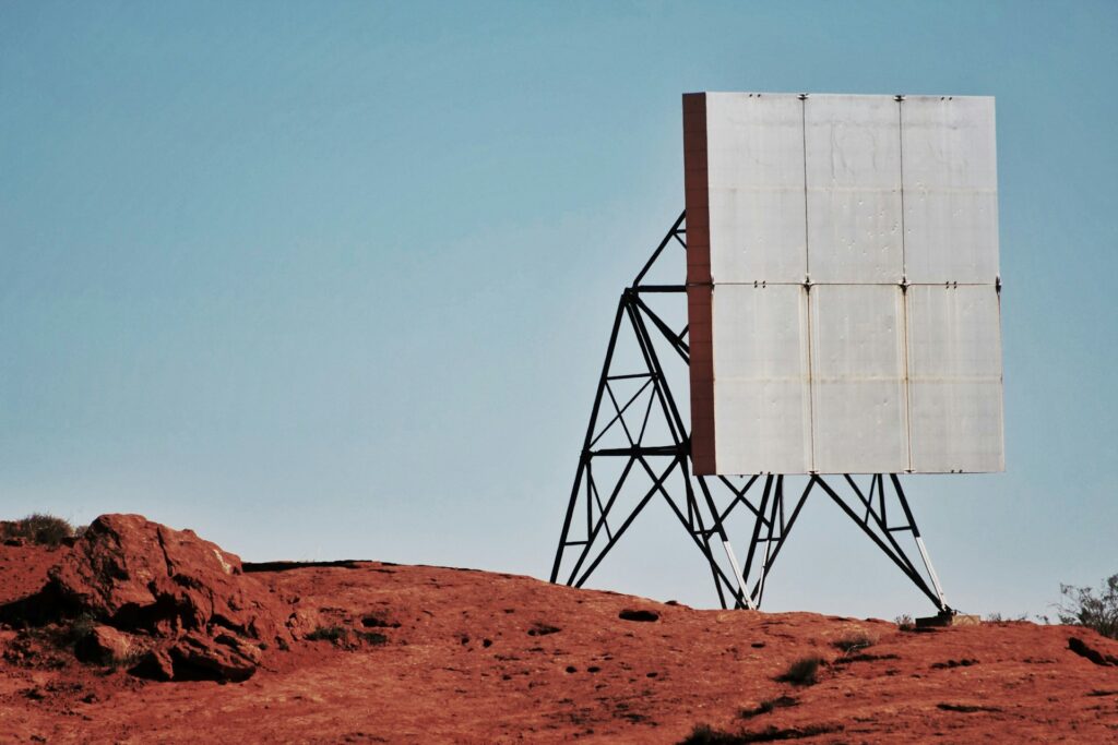 A block of solar panels, possibly hydropanels, sits on a red rocky outcropping in a desert-like environment. The sky is cloudless and blue above it.