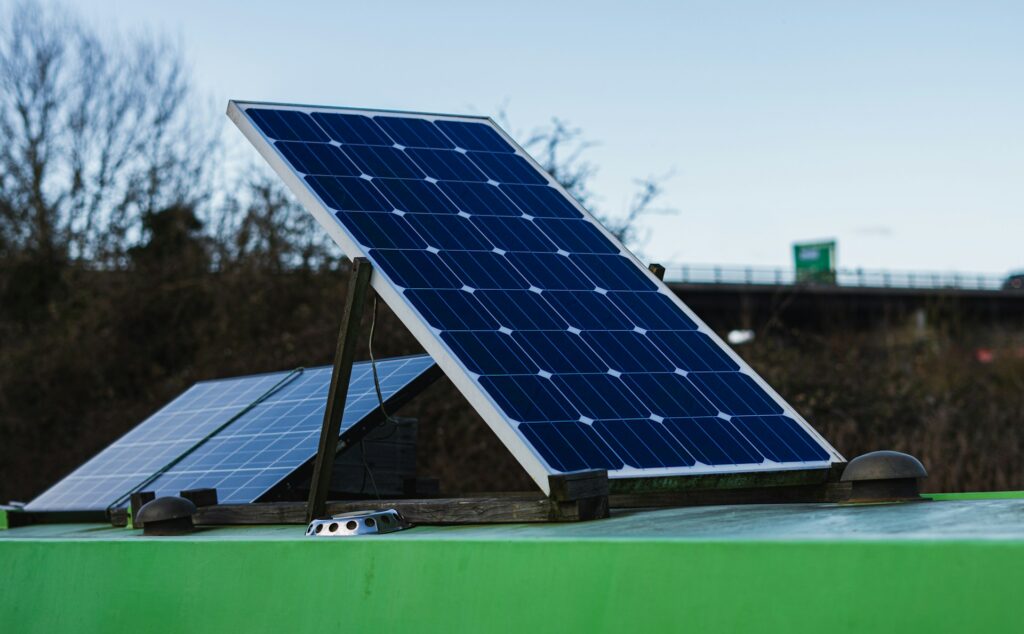 A small solar panel sits on top of a green structure on the side of an interstate. The interstate's bridge and some trees in the dead of winter are in the background under a blue sky.