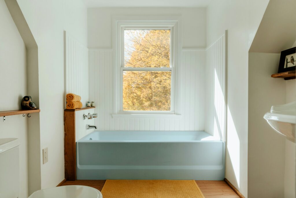 A light blue tub sits at the end of a bathroom within three walls covered in white shiplap boards. A small window is above the tub. The other walls are white and there's a white stand-alone porcelain sink to the right.
