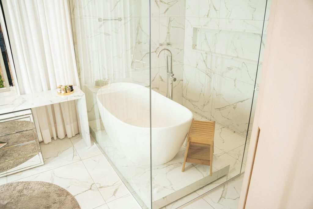 A white stand-alone tub sits on a marble floor encased in glass walls. A wooden bench stands next to it. A marble counter is off to the side with glass drawers beneath.