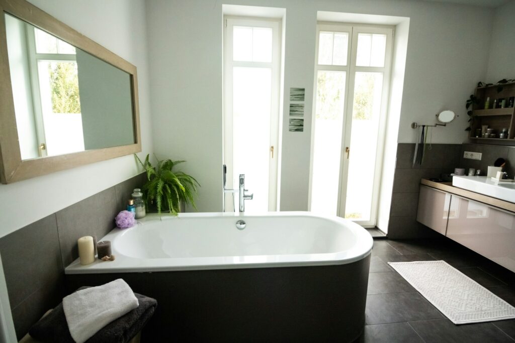 A white bathtub sits in a dark bathroom with pale green paint. Two glass doors are in the wall on one end. A mirror stretches across the other wall behind the tub.