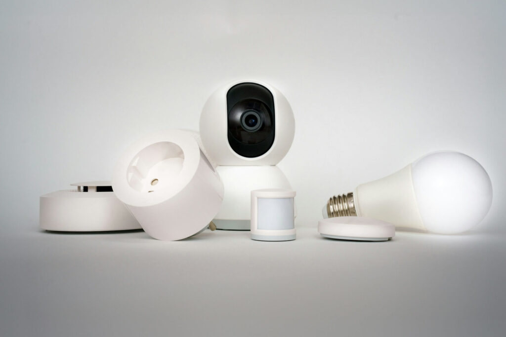 A white standing home security camera, a light bulb, two sensors, a smart plug and an alarm sit against a white background.