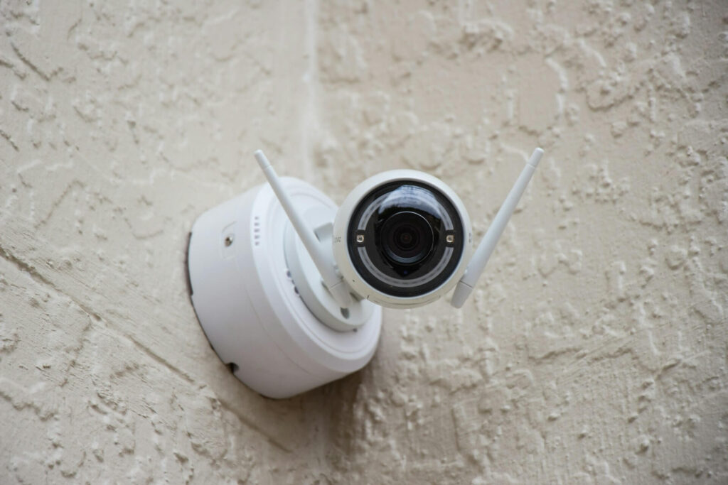 A white outdoor camera with two white antennae is mounted in the corner of two white stucco walls. It's connected to one of the more popular home security camera systems.