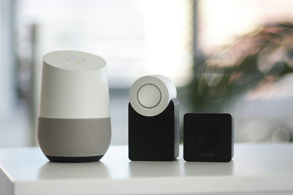 a Nuki security sensor, a stand and a control unit that's white and gray sit on a white counter. A window and the large branch of an indoor plant are blurry in the background.