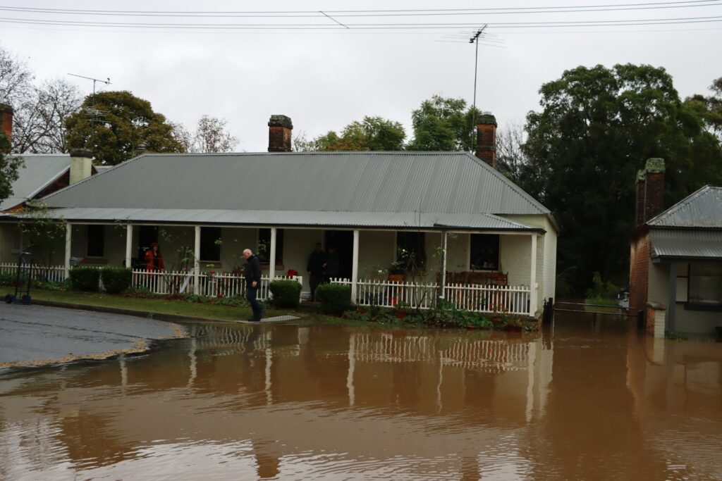 A one-story house with a gray roof and a small strip of landscaping in front of it sits in a flood of brown water. An older man in a black jacket steps outside of the house and looks at the water while two people watch from the porch. The food is one of the things that damages foundations.