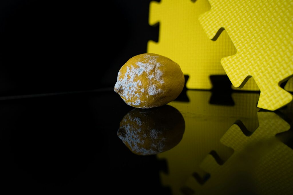 An uncut lemon covered in white mold sits on a glassy black surface with two large yellow foam puzzle pieces standing vertically in the background.