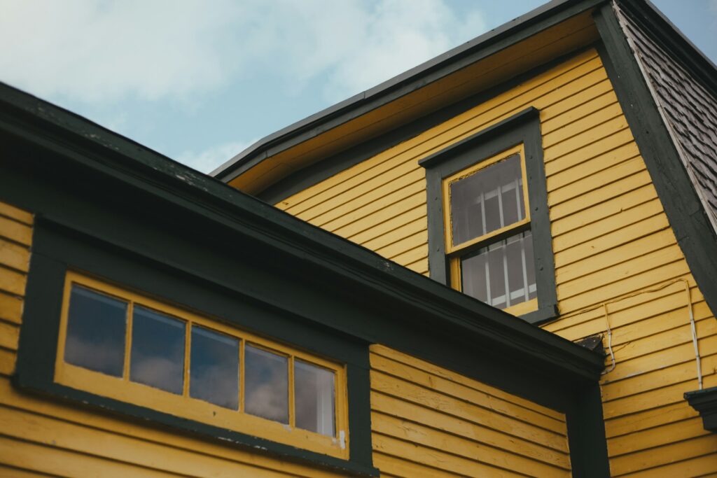 A bright yellow house with gray trim sits under a cloudy blue sky. Its gutters are older and potentially clogged.