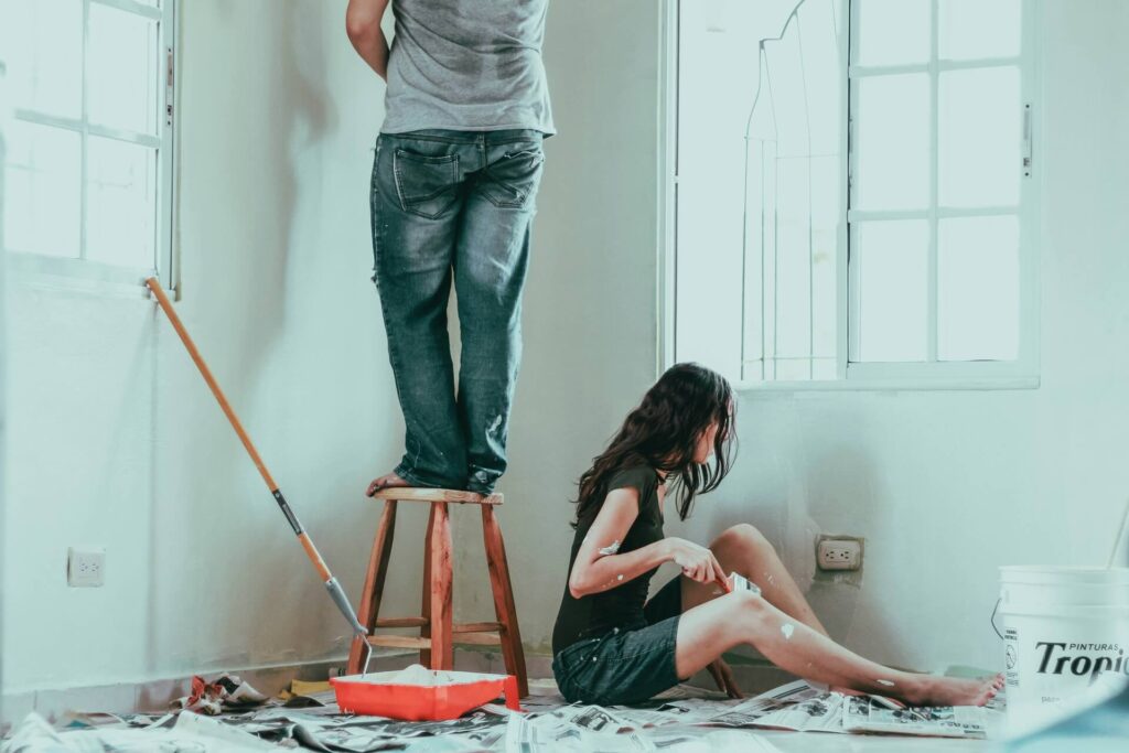 A couple painting a room surrounded by materials they need for the job. 