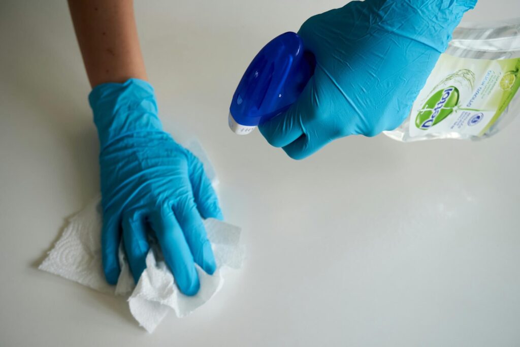 A person wearing rubber gloves using a detergent and paper towel to clean a surface